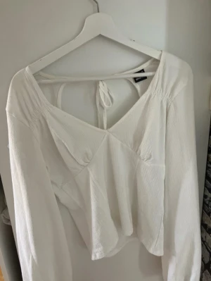 Vit långärmad blus med knyt i ryggen - Säljer en vit blus med långa ärmar och v-ringning. Blusen har en snygg detalj med knyt i ryggen och lätt strukturerat tyg som ger en avslappnad men trendig look. Har en liten fläck på ryggen, men den syns knappast!