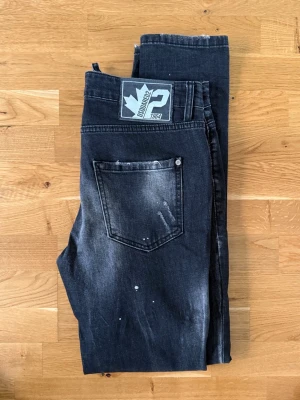 D2 jeans med slitningar - Säljer ett par svarta jeans med coola slitningar och blekta detaljer. Jeansen har en klassisk femficksmodell och vita färgstänk för en trendig look. Passar perfekt till dig som gillar streetwear och vill ha en avslappnad stil.