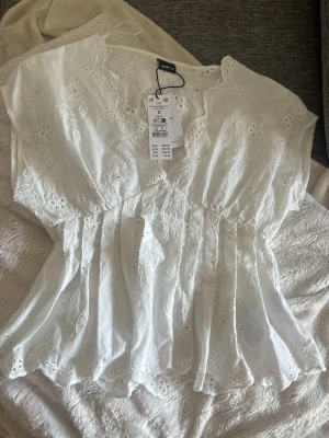 Vit broderad blus från gina - Söt vit blus med broderade blommor och spetsdetaljer. Blusen har en markerad midja med smock baktill och vacker vågig kant nertill. Kortärmad modell i luftig bomull, perfekt för sommardagar. Romantisk och feminin stil. Prislapp kvar, endast provad annars oanvänd🥰