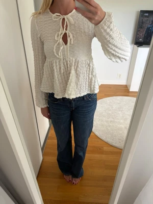 Vit blus med knyt och volang - Supersöt vit blus med lång ärm, struktur i tyget och två knyt framtill. Blusen har volang nedtill och en lätt v-ringad halsringning. Man kan även gömma det översta snöret och göra den mer urringad. Tröjan passar även xs