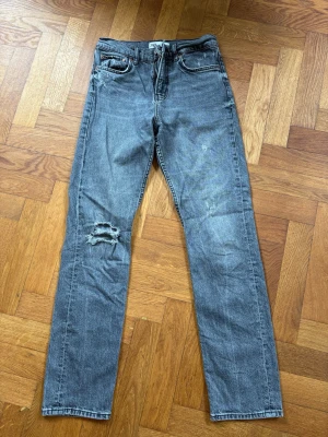 Grå raka jeans från ZARA - Snygga grå jeans från ZARA med raka ben och slitning på vänster knä. Klassisk femficksmodell med dragkedja och knapp. Perfekta för en avslappnad och trendig look.