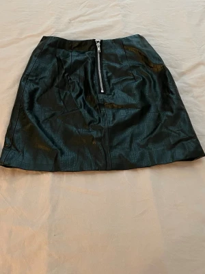 Svart kortkjol i skinnimitation H&M - Säljer en svart kortkjol från H&M Divided i skinnimitation med croc-mönster och dragkedja framtill. Kjolen har en cool look och är fodrad med polyester. Perfekt för dig som vill ha en edgy vibe till outfiten.