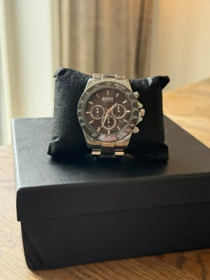 Silverfärgad klocka  - Stilren klocka  med silverfärgad länkarmband och svart urtavla. Klockan har flera små urtavlor, tydliga markeringar och kronograf-funktion. Perfekt accessoar för dig som gillar en klassisk och modern look.