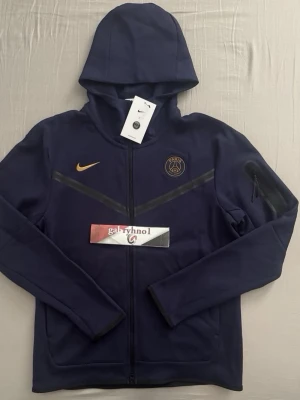 Mörkblå PSG hoodie med dragkedja - Snygg mörkblå hoodie från Nike med PSG-logga på bröstet och guldfärgad Nike-swoosh. Hoodien har hel dragkedja, huva och diskreta svarta detaljer över bröstet. Perfekt för dig som gillar sportig stil och vill representera Paris Saint-Germain. Inga tags