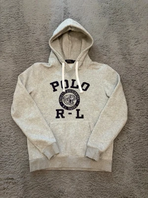 Grå hoodie från Polo Ralph Lauren - Snygg grå hoodie från Polo Ralph Lauren med svart tryck framtill där det står 'POLO R-L' och en tigerlogga. Klassisk huva med vita snören och magficka. Perfekt för en avslappnad och sportig stil. ⚠️⚠️tröjan var i storlek M men har krympt till storlek S⚠️⚠️