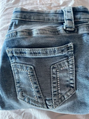 Ljusblå bootcut jeans med strass - Säljer ett par ljusblå bootcut jeans med snygga strassdetaljer på bakfickorna. Jeansen har klassisk femficksmodell, normal midja och är i mjukt denimtyg. Perfekta för dig som vill ha en trendig och bekväm look.