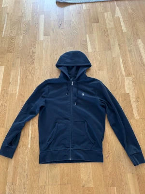 Marinblå hoodie från Polo Ralph Lauren - Snygg marinblå hoodie med dragkedja från Polo Ralph Lauren. Klassisk design med huva, två fickor framtill och den ikoniska broderade loggan på bröstet. Perfekt för en avslappnad och stilren look. Tillverkad i mjukt bomullsmaterial.