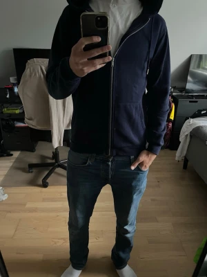 Mörkblå zip-hoodie med huva - En stilren mörkblå hoodie med dragkedja framtill och två fickor. Hoodien har en klassisk huva och ribbade muddar vid ärmslut och nederkant. Perfekt för en avslappnad look och enkel att matcha med jeans eller joggers. Nopprig!