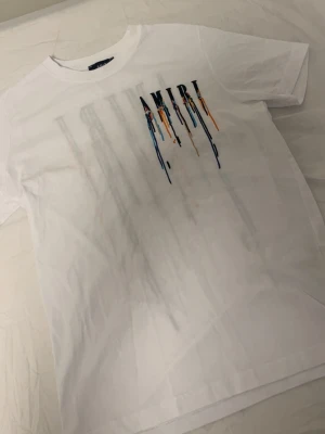 Vit AMIRI t-shirt med färgstänk - Säljer en vit t-shirt från AMIRI med coolt tryck framtill. T-shirten har färgstänk i blått, orange och svart som rinner ner från loggan. Klassisk passform och rund halsringning. Perfekt för dig som gillar streetwear och vill sticka ut.
