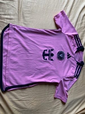 Rosa Inter Miami Messi matchtröja - Säljer en rosa Inter Miami matchtröja från Adidas med Messi och nummer 10 på ryggen. Tröjan är rosa och har svarta detaljer, korta ärmar och klubbmärke på bröstet. Perfekt för dig som vill stötta laget och Messi på match eller träning.