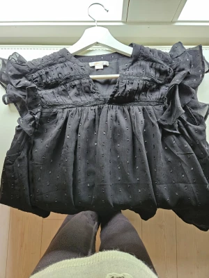 Svart blus - Supersöt svart blus från Gina Tricot med volangdetaljer på axlarna och små prickar i tyget. Blusen har v-ringning och är gjord i ett lätt, luftigt material som känns skönt mot huden. Perfekt för dig som gillar en romantisk och trendig stil.