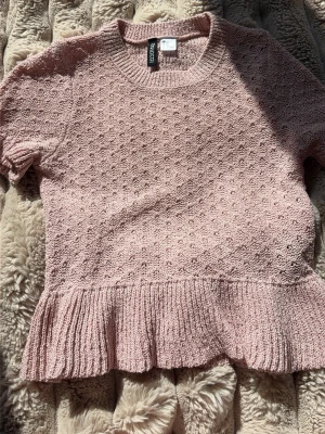 Ljusrosa stickad topp med volang - Supersöt ljusrosa stickad topp med korta ärmar och volangkant nedtill. Tröjan har ett diskret hålmönster över hela och rund halsringning. Perfekt för dig som gillar en feminin och trendig stil.