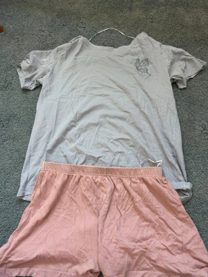 Ljusgrå pyjamaströja & rosa shorts - Tvådelad pyjamas med en ljusgrå t-shirt och ljusrosa shorts. T-shirten har korta ärmar och ett litet tryck med fjäril på bröstet. Shortsen har resår i midjan och är gjorda i mjukt bomullstyg. Perfekt för mysiga hemmakvällar.