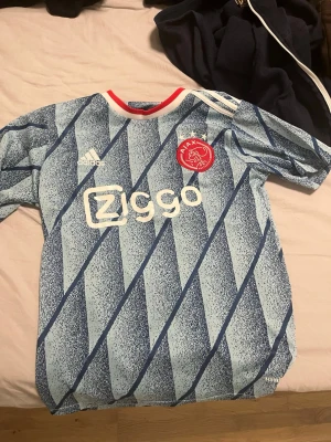 Ajax blå matchtröja från Adidas - Snygg blå Ajax matchtröja från Adidas med diagonala mönster i olika blå nyanser. Tröjan har vita detaljer, röd krage och Ajax klubbmärke på bröstet samt Ziggo-logga framtill. Perfekt för fotbollsträning eller att bära till vardags.