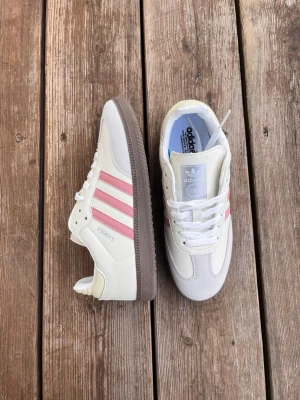Adidas Samba vita med rosa ränder - Säljer ett par Adidas Samba sneakers i vitt med rosa ränder och brun sula. Klassisk låg modell med ovandel i läder och mockadetaljer. Ikonisk retrostil som passar perfekt till streetwear och casual outfits. 