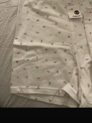 Två vita pyjamasshorts XS Kappahl - Säljer två par vita pyjamasshorts från Kappahl i storlek XS. Ena paret har små röda hjärtan och det andra har ett gulligt blommönster i rosa och grönt. Båda har hög midja och är gjorda i mjukt bomullsmaterial. Perfekta för mysiga hemmakvällar.