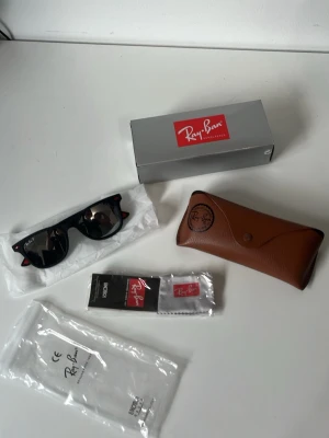 Svarta Ray-Ban Wayfarer solglasögon - Klassiska svarta Ray-Ban Wayfarer solglasögon med ikonisk form. Kommer med originalbrun fodral, putsduk och box. Snygg och tidlös design som passar till allt. Perfekt accessoar för soliga dagar.