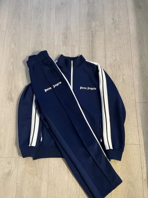 Palm Angels tracksuit S - Säljer en mörkblå tracksuit från Palm Angels med vita ränder längs ärmar och ben. Setet består av zip-jacka och byxor med resår i midjan och muddar vid ärmslut. Palm Angels-logga broderad på både jacka och byxa. Perfekt för en sportig och trendig look.