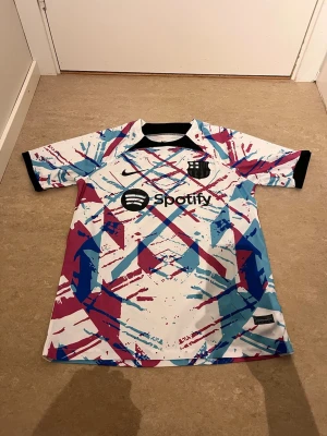 FC Barcelona Nike matchtröja färgstark - Säljer en FC Barcelona matchtröja från Nike med färgstarkt grafiskt mönster i vitt, blått, rött och svart. Tröjan har korta ärmar, svart krage och klubbmärke samt Spotify-logga på bröstet. Perfekt för dig som älskar fotboll och vill sticka ut på planen.