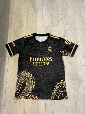 Real Madrid T-shirt S - Snygg svart Real Madrid matchtröja med guldiga drakdetaljer och tryck. Framsidan har Emirates Fly Better-logga och klubbmärke, baksidan har Mbappé och nummer 9 i guld. Korta ärmar och rund hals. Perfekt för fotbollsfans!