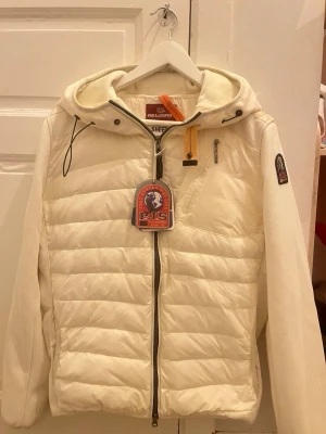Vit pufferjacka från Parajumpers - Snygg vit pufferjacka från Parajumpers med huva och dragkedja framtill. Jackan har quiltad design, ribbade ärmar och en bröstficka med dragkedja. Märkets patch sitter på ärmen och det finns en gul detalj vid halsen. Perfekt för kalla dagar.Priset går att diskutera vid snabb affär, finn de frågor så är de bara o skriv 😊
