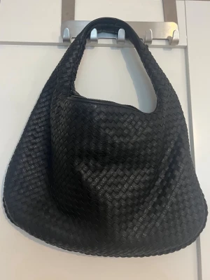 Svart flätad väska - Svart axelväska i flätad design med mjuk, rundad form från Design by si. Väskan har dragkedja upptill och ett rymligt innerfack med extra fickor.