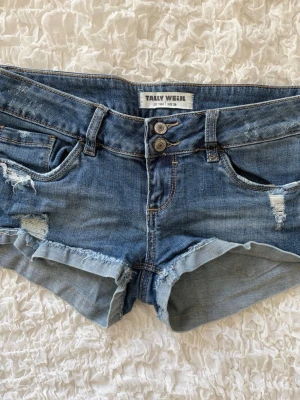 Snygga slitna jeansshorts från Tally Weijl - Superfina och trendiga lågmidjade jeansshorts i en klassisk blå tvätt från Tally Weijl. Shortsen har snygga slitna detaljer både fram och bak, samt uppvikta benslut. Knäpps med två knappar och dragkedja fram. Storlek 36. Midjemåttet rakt över är 37cm (alla mått finns i bilderna). Skickas snabbt!!
