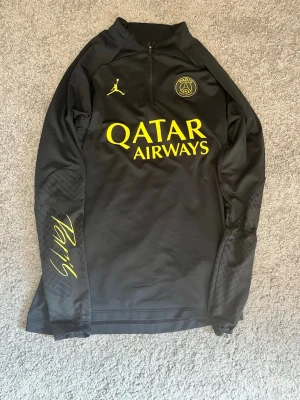 Psg tröja - Säljer en svart långärmad träningströja från PSG x Jordan med gul text och logga. Tröjan har halv dragkedja, trycket 'Qatar Airways' på bröstet och 'Paris' på ärmen. Tillverkad i ett sportigt, stretchigt material som passar perfekt till träning.