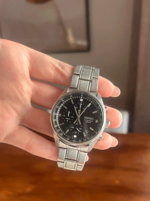 Seiko herrklocka i silver med svart urtavla - Stilren Seiko herrklocka med svart urtavla och silverfärgat armband i metall. Klockan behöver byta batteri och har lite ripor på glaset annars funkar den som den ska😇