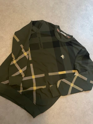 Burberry  - Snygg grön tröja från Burberry med klassiskt rutmönster i svart, gult och beige. Tröjan har rund halsringning och ribbade muddar. Ikonisk Burberry-logga på bröstet och tryckt märke på ärmen. Perfekt för dig som gillar streetstyle.