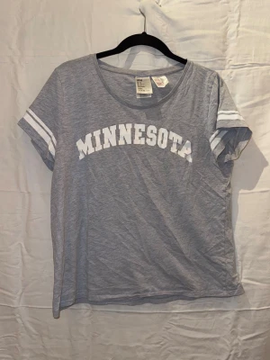 Grå t-shirt med Minnesota-tryck - Snygg grå t-shirt med vit text 'MINNESOTA' över bröstet och vita ränder på ärmarna. T-shirten har rund halsringning och korta ärmar. Perfekt för en avslappnad och sportig look.