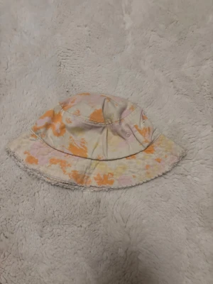 Flerfärgad solhatt - Säljer en solhatt med tie-dye mönster i orange, ljusrosa och gul. Hatten har bred brätte och små ventilationshål på sidorna. Perfekt accessoar för en avslappnad och trendig stil.
