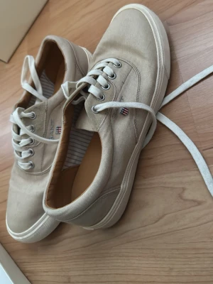 Beige sneakers från Gant - Snygga beige sneakers från Gant med klassisk låg profil och vita snören. Skorna har ovandel i canvas och diskret logga på sidan. Perfekta för en stilren och avslappnad look. Kanske använda max en gång stått i garderoben i över 5 år aldrig använts.