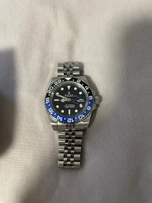  GMT-Master II silver/blå klocka - Snygg  GMT-Master II klocka med silverfärgad länk i rostfritt stål och svart urtavla. Bezel i svart och blått med 24-timmarsmarkeringar. Klockan har tydliga index och datumfönster vid 3. Perfekt accessoar för dig som gillar klassisk och sportig stil. OBS helt ny du får med allt på bilden och för att kunna justera till din storlek.