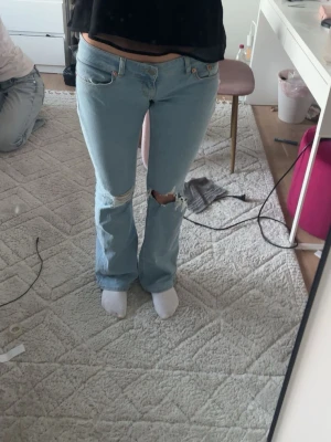 Ljusblå bootcut jeans med hål - Säljer ett par ljusblå bootcut jeans med slitna detaljer och hål på ena knät. Jeansen har klassisk femficksdesign och låg midja. Perfekta för en avslappnad och trendig look.
