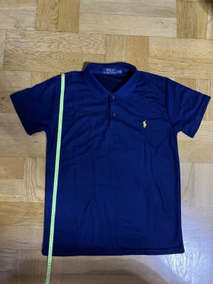 Marinblå pikétröja från Polo Ralph Lauren - Snygg marinblå pikétröja från Polo Ralph Lauren med klassisk krage och två knappar. Ikoniska gula loggan broderad på bröstet. Tillverkad i mjuk bomull som känns skön mot huden. Perfekt till jeans eller shorts för en clean look.
