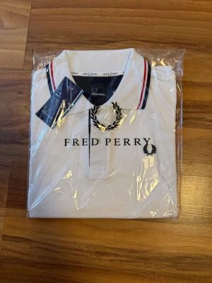 Vit Fred Perry pikétröja - Snygg vit pikétröja från Fred Perry med klassisk krage och rödblå ränder på kragen. Tröjan har broderad logga på bröstet och knappar framtill. Perfekt för en stilren och sportig look.