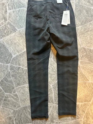 Mörkgrå rutiga kostymbyxor Jack & Jones - Snygga mörkgrå kostymbyxor från Jack & Jones med diskret rutmönster. Byxorna har två bakfickor med knapp, bälteshällor och smal passform. Perfekta för en stilren look och lätta att matcha med skjorta eller t-shirt.