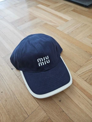 Mörkblå keps från Miu Miu - Snygg mörkblå keps från Miu Miu med vit broderad logga framtill och vit kant runt skärmen. Klassisk form och tillverkad i bomull, perfekt för en trendig och avslappnad look.