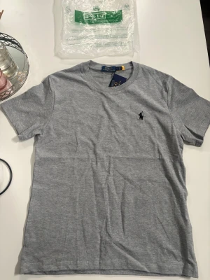 Grå t-shirt från Polo Ralph Lauren - Snygg grå t-shirt från Polo Ralph Lauren med klassisk rund halsringning och den ikoniska svarta logobroderingen på bröstet. Tillverkad i mjuk bomull för en skön känsla. Perfekt basplagg till garderoben.