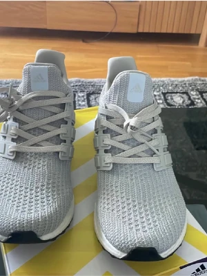Grå Adidas Ultraboost sneakers - Snygga grå Adidas Ultraboost sneakers med stickad ovandel i mesh och klassiska tre ränder på sidan. Skorna har vita detaljer och en bekväm, responsiv sula. Perfekta för dig som gillar stilrena och sportiga sneakers. OBS BOXEN FINNS INTE 