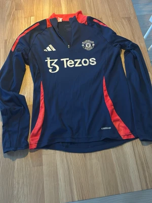 Manchester United träningströja blå/röd - Snygg långärmad träningströja från Manchester United i marinblått med röda detaljer och halv dragkedja. Tröjan har klubbmärke och Adidas-logga på bröstet samt Tezos-tryck framtill. Tillverkad i funktionsmaterial (AEROREADY) som andas.