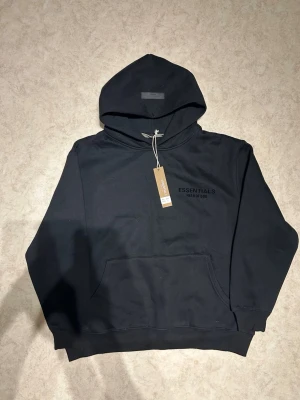 Svart Essentials Fear of God hoodie - Snygg svart hoodie från Essentials Fear of God med diskret logga på bröstet. Klassisk känguruficka framtill och huva. Tillverkad i mjukt bomullsmaterial som känns skönt mot huden. Perfekt för en avslappnad och trendig streetwear-look.