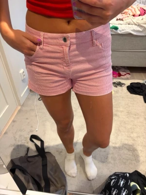 Rosa och vita randiga shorts - Säljer ett par snygga shorts med rosa och vita smala ränder. De har hög midja, klassisk fickdesign och stängs med knapp och dragkedja. Perfekta för sommaren och tillverkade i mjuk bomull för skön känsla.