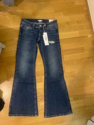 Blå bootcut jeans med broderade fickor - Säljer ett par blå bootcut jeans i denim med slitning på låret och snygga broderade detaljer på bakfickorna. Jeansen har klassisk femficksmodell, normal midja och lätt utsvängda ben för en retro känsla.