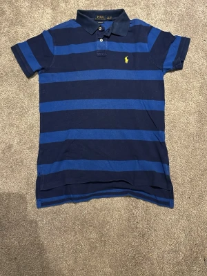Randig pikétröja från Polo Ralph Lauren - Mörkblå och blå randig pikétröja med klassisk krage och korta ärmar. Ikoniska Polo Ralph Lauren-loggan broderad på bröstet i gult. Perfekt till jeans eller shorts för en avslappnad stil.