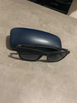 Svarta solglasögon från Lacoste - NYPRIS:1100KR   Mitt pris 150kr.   Snygga svarta solglasögon från Lacoste med klassisk rektangulär form. Glasögonen har mörka linser och stilren design, perfekt för att lyfta din look. Kommer med ett blått fodral med Lacoste-logga. Ena sidan är väldigt enkel att dra fram och tillbaka men inget som påverkar produkten 