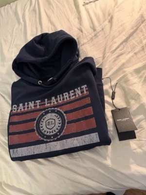 Saint Laurent hoodie - Säljer en mörkblå hoodie från Saint Laurent med tryck i rött, vitt och blått framtill. Hoodien har huva med dragsko och en stor magficka. Materialet ser ut att vara mjuk bomull, perfekt för en avslappnad och cool stil.