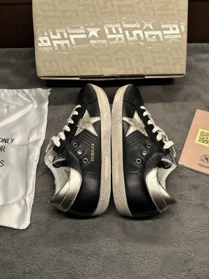 Svart/silver golden goose superstars - Snygga svarta sneakers med vit sula och silverfärgade detaljer, inklusive en stor stjärna på sidan och metallic häl. Skorna har vita skosnören och en klassisk låg profil. Perfekta för dig som vill sticka ut med en cool och trendig look.