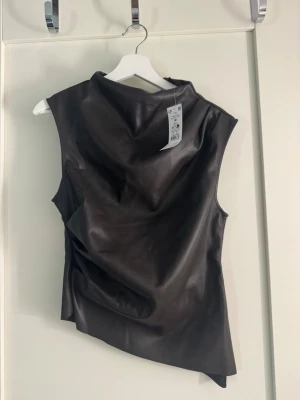 Svart asymmetrisk ärmlös skinn-topp - Ärmlös draperad topp i skinnimitation från Gina Tricot i storlek M. Materialet är skönt på insidan och stretchigt. Sitter fint på och formar kroppen bra. Inte kommit till användning tyvärr då jag har för mycket kläder, därav prislappen kvar!